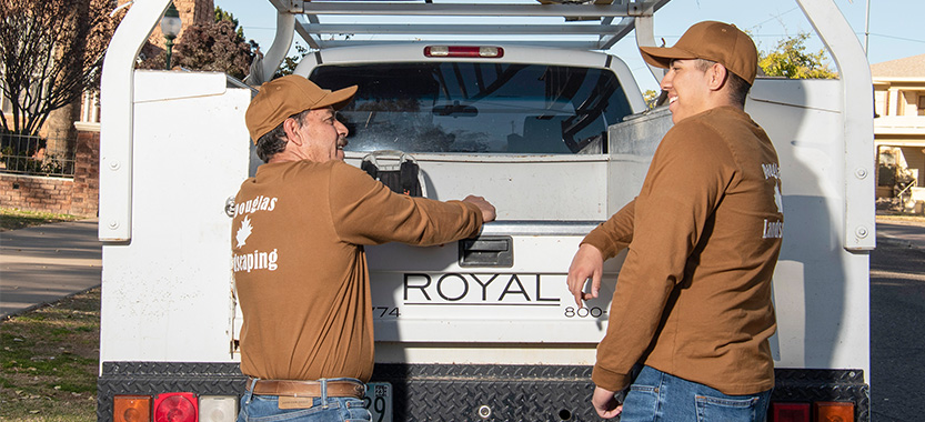 two workers stand behind their white landscaping truck